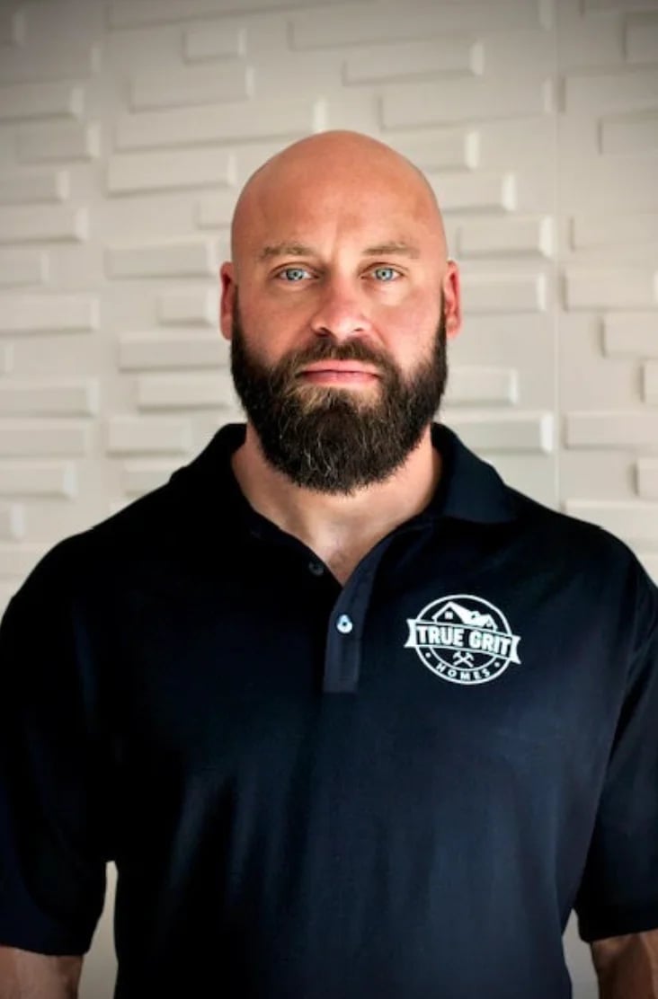 Bald man with full dark beard wearing dark navy polo shirt with True Grit logo, photographed against white brick wall background