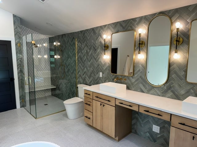 Modern bathroom with herringbone tile walls, double vanity with mirrors, walk-in shower, and brass fixtures